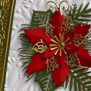 May include: A red poinsettia flower with gold accents and green leaves. The flower is on a white background with a gold and green patterned border.