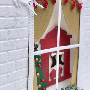 May include: A white brick wall with a window cut out. The window has a gold curtain and a red fireplace with stockings hanging from it. There is a green Christmas tree and a red cardinal on a branch outside the window.