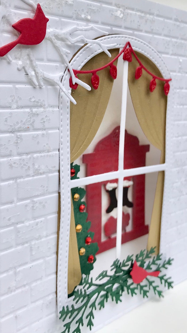 May include: A white brick wall with a window cut out. The window has a gold curtain and a red fireplace with stockings hanging from it. There is a green Christmas tree and a red cardinal on a branch outside the window.