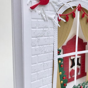 May include: A white brick wall with a window cut out. The window has a red curtain and a view of a fireplace with stockings hanging from it. A green Christmas tree is in the window with gold and red ornaments. A cardinal bird is perched on a branch above the window.