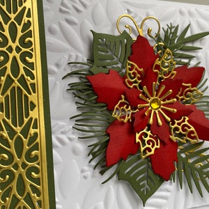 May include: A white card with a red poinsettia flower, green leaves, and gold accents. The card has a textured background and a gold border.