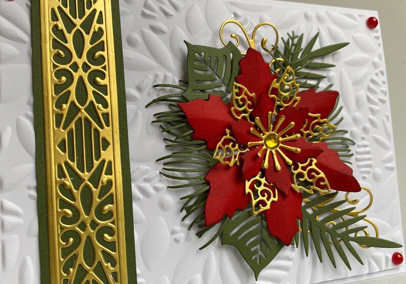 May include: A white card with a red poinsettia flower, green leaves, and gold accents. The card has a textured background and a gold border.