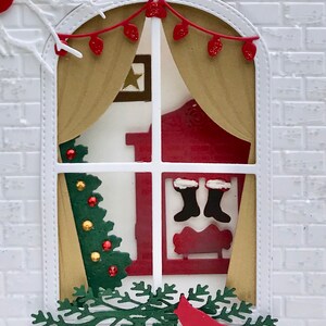 May include: A white brick house with a window decorated for Christmas. The window has a red and gold curtain, a small Christmas tree, and a fireplace with stockings hanging from it. There is a red cardinal perched on a green branch below the window.