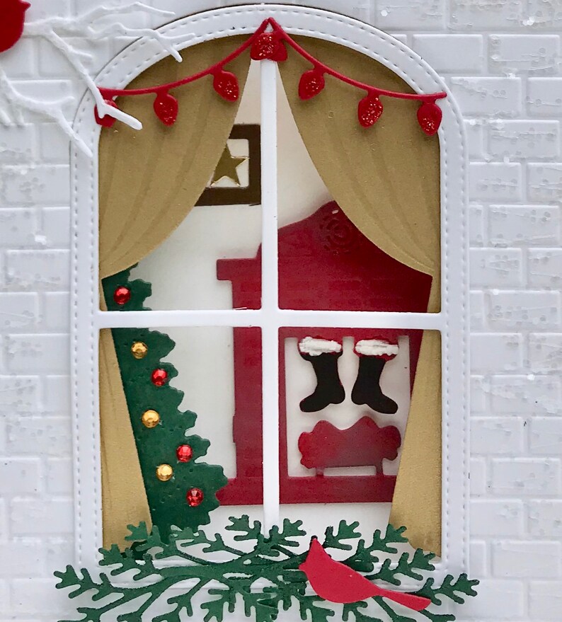 May include: A white brick house with a window decorated for Christmas. The window has a red and gold curtain, a small Christmas tree, and a fireplace with stockings hanging from it. There is a red cardinal perched on a green branch below the window.