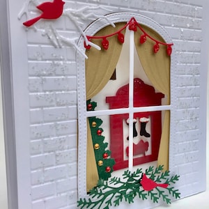 May include: A white brick wall with a window featuring a red fireplace, stockings, and a Christmas tree. The window is decorated with gold curtains and red lights. A red cardinal is perched on a branch outside the window.