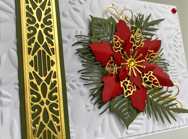 May include: A handmade Christmas card featuring a red poinsettia with gold accents and green foliage. The card has a white background with an embossed pattern and a gold and green border.
