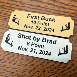 May include: Two gold and silver metal plaques with black text and antler silhouettes. The top plaque reads "First Buck 10 Point Nov. 22, 2024". The bottom plaque reads "Shot by Brad 8 Point Nov. 21, 2024".