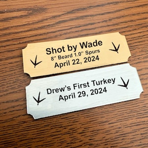 May include: Two gold and silver metal plaques with black text. The top plaque reads "Shot by Wade 8" Beard 1.0" Spurs April 22, 2024". The bottom plaque reads "Drew's First Turkey April 29, 2024".
