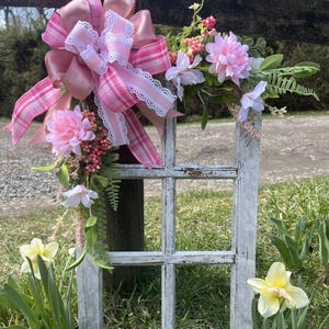 May include: A white, weathered window frame with a pink and white plaid bow and floral accents. The window frame is surrounded by green grass and yellow daffodils.
