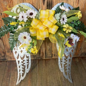 May include: A pair of metal angel wings decorated with a yellow and white checkered bow, yellow mesh, and white and yellow flowers. A small photo is attached to the right wing.