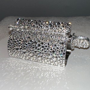 Puede incluir: Un bolso de mano plateado, rectangular, cubierto de brillantes pedrería. El bolso tiene una tapa con bisagras y un asa pequeña. La superficie está completamente cubierta de pedrería, lo que le da un aspecto glamuroso.