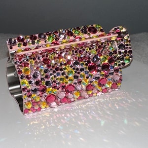 Puede incluir: Un portarrollos de papel higiénico adornado con pedrería multicolor. El soporte está cubierto con una variedad de pedrería rosa, morada, amarilla y transparente. El soporte tiene una base de metal plateado y un asa curvada.