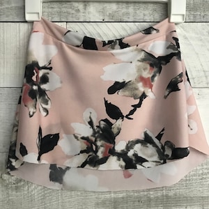 May include: A pale pink skirt with a floral design. The skirt displays large white, black, and grey flowers. It hangs on a white hanger against a light wood backdrop. The skirt has a layered design.