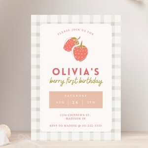 May include: A light pink and cream-colored invitation card with a gingham border. The card features the text "OLIVIA'S berry first birthday" with a strawberry illustration. The date and time are also included.