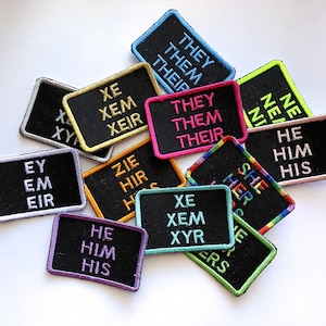 May include: A collection of colorful embroidered patches with various pronoun sets, including "they/them/their", "he/him/his", "she/her/hers", "xe/xem/xeir", "ey/em/eir", and "zie/hir/his".