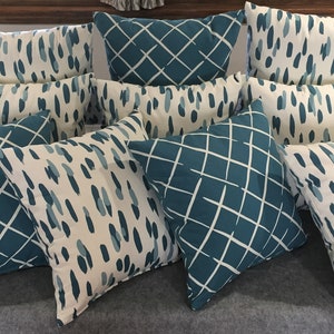 May include: A collection of nine decorative throw pillows with a teal and white geometric pattern. The pillows are arranged in a staggered pattern on a gray surface.