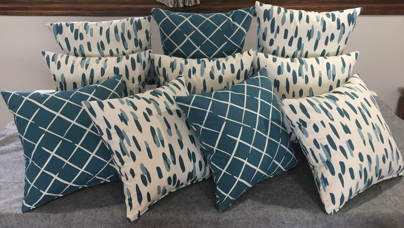 May include: A collection of nine decorative throw pillows with a teal and white geometric pattern. The pillows are arranged in a staggered pattern on a gray surface.