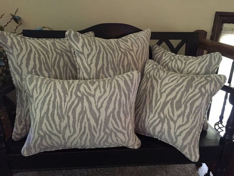 Op de afbeelding: Vijf decoratieve kussens met een zebraprint in grijstinten en wit. De kussens zijn gerangschikt op een zwarte bank.