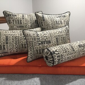 Op de afbeelding: Vijf kussens en een rolkussen met een beige stof en zwarte tekst. De kussens hebben een patroon van stadsnamen in zwarte tekst op een beige achtergrond. De kussens zijn gerangschikt op een oranje bank.
