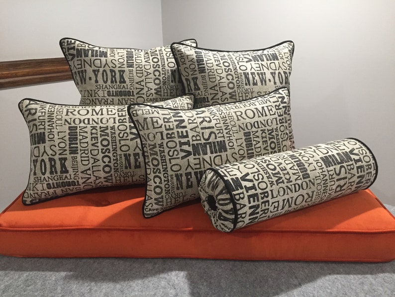 Op de afbeelding: Vijf kussens en een rolkussen met een beige stof en zwarte tekst. De kussens hebben een patroon van stadsnamen in zwarte tekst op een beige achtergrond. De kussens zijn gerangschikt op een oranje bank.