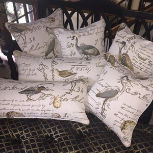 Op de afbeelding: Een set van zes decoratieve kussens met een cr&egrave;mekleurige achtergrond en bruine schrijftekst. De kussens bevatten verschillende vogelillustraties, waaronder reigers, een kleine vogel en een ei. De kussens zijn gerangschikt op een zwart en gouden gepatenteerd oppervlak.
