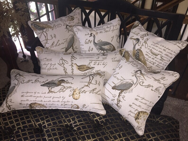 Op de afbeelding: Een set van zes decoratieve kussens met een cr&egrave;mekleurige achtergrond en bruine schrijftekst. De kussens bevatten verschillende vogelillustraties, waaronder reigers, een kleine vogel en een ei. De kussens zijn gerangschikt op een zwart en gouden gepatenteerd oppervlak.