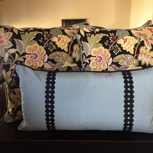 May include: Three decorative pillows on a wooden bench. Two pillows have a black background with a floral pattern in shades of red, yellow, and green. The third pillow is light blue with black trim and a scalloped design.
