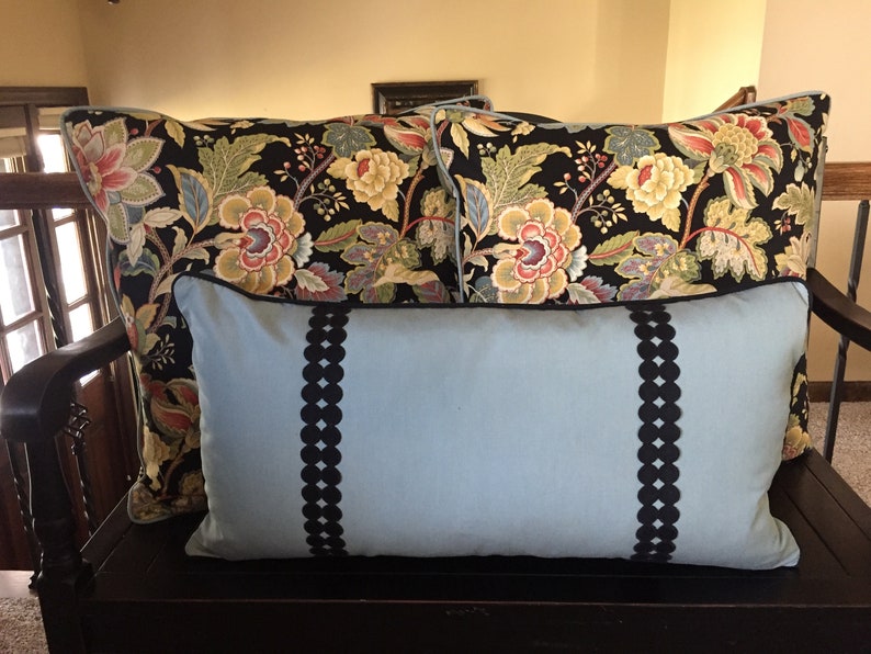 May include: Three decorative pillows on a wooden bench. Two pillows have a black background with a floral pattern in shades of red, yellow, and green. The third pillow is light blue with black trim and a scalloped design.