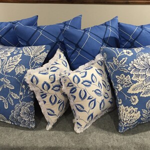 Op de afbeelding: Een verzameling blauwe en witte gepatroneerde kussens met verschillende bloemen- en geometrische ontwerpen. De kussens zijn gerangschikt op een grijs oppervlak.