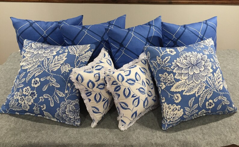 Op de afbeelding: Een verzameling blauwe en witte gepatroneerde kussens met verschillende bloemen- en geometrische ontwerpen. De kussens zijn gerangschikt op een grijs oppervlak.