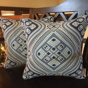 Op de afbeelding: Twee decoratieve kussens met een geometrisch patroon in tinten blauw, grijs en beige. De kussens hebben een donkerblauwe achtergrond met een lichtblauw ruitpatroon en een witte rand.