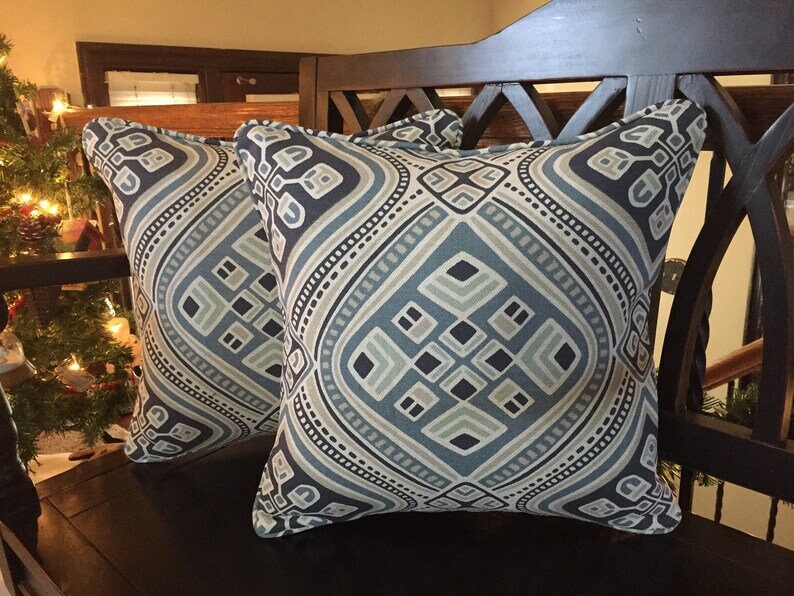 Op de afbeelding: Twee decoratieve kussens met een geometrisch patroon in tinten blauw, grijs en beige. De kussens hebben een donkerblauwe achtergrond met een lichtblauw ruitpatroon en een witte rand.