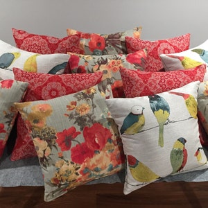May include: A collection of decorative throw pillows featuring floral and bird patterns. The pillows are in shades of red, green, yellow, and blue, with a white background.
