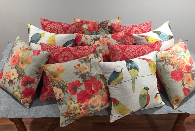 May include: A collection of decorative throw pillows featuring floral and bird patterns. The pillows are in shades of red, green, yellow, and blue, with a white background.