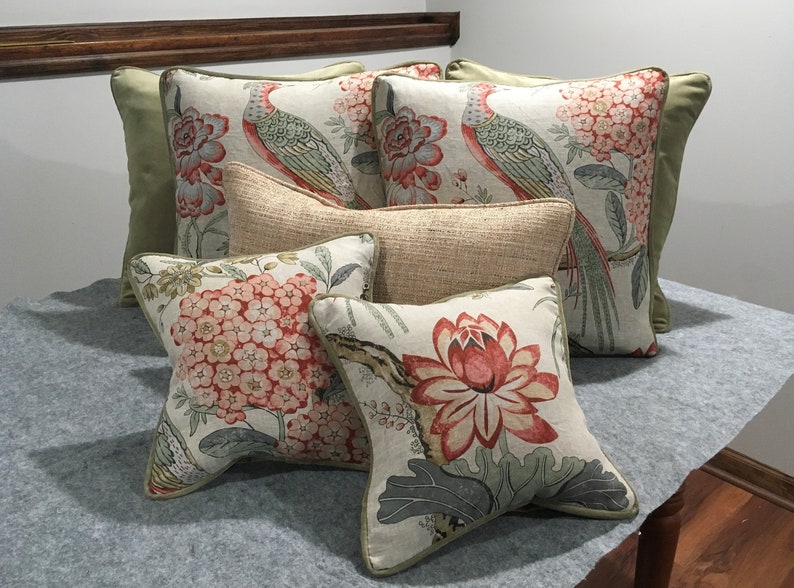 May include: Five decorative throw pillows with floral and bird patterns. The pillows are arranged on a light gray surface. The pillows are in shades of green, red, and white.