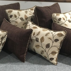 May include: A set of eight decorative throw pillows featuring a brown and cream leaf pattern. The pillows are rectangular and have a brown fabric with a subtle texture. The leaf pattern is embroidered in cream and brown thread. The pillows have a brown trim around the edges.