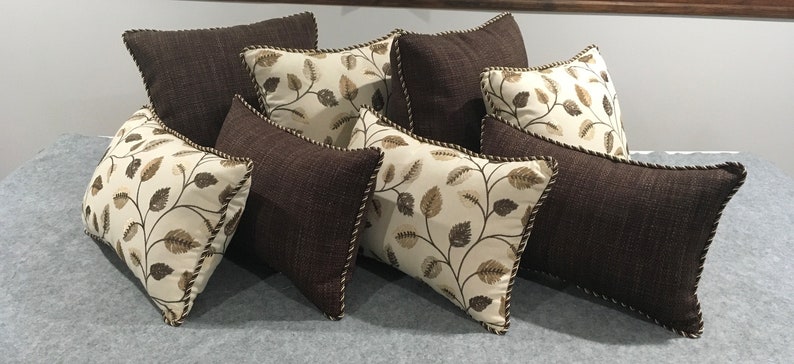 May include: A set of eight decorative throw pillows featuring a brown and cream leaf pattern. The pillows are rectangular and have a brown fabric with a subtle texture. The leaf pattern is embroidered in cream and brown thread. The pillows have a brown trim around the edges.