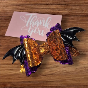 Op de afbeelding: Een haarstrik met Halloween-thema en vleermuisvleugels. De strik heeft gouden en paarse pailletten, met zwarte vleermuisvleugels aan elke kant. Een roze kaart met de tekst "Thank you" staat op de achtergrond.
