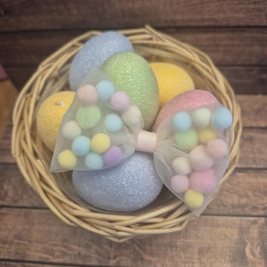 May include: A pastel-colored Easter egg bow with a tulle bow and colorful pom-poms. The bow is sitting in a wicker basket with pastel-colored Easter eggs.