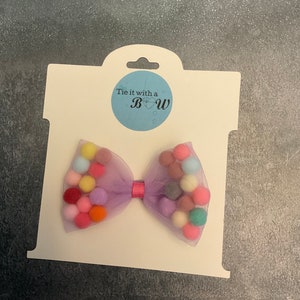 May include: A purple tulle bow with a pink ribbon in the center. The bow is decorated with colorful pom-poms. The bow is on a white card with the text "Tie it with a Bow" in a blue circle.