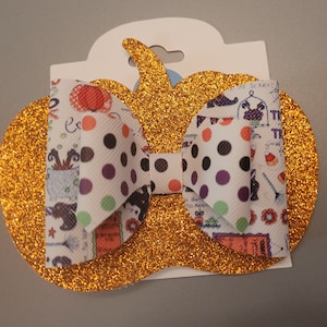 Op de afbeelding: Een gouden glitter-pompoenvormige haarstrik met een witte strik met zwarte en paarse stippen. De strik heeft een Halloween-thema print met afbeeldingen van heksen, katten en pompoenen.