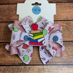 Può includere: Un fiocco di glitter rosa e bianco con un tema di ritorno a scuola. Il fiocco presenta una pila di libri, una mela, una lampadina e le parole "ABC", "123", "Ritorno a scuola" e "Amore".