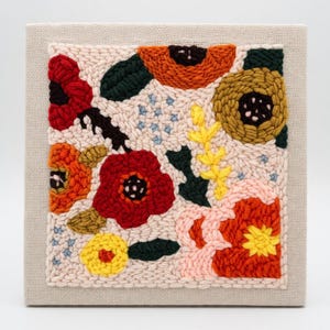 May include: A square, framed textile art piece featuring a colorful floral design. The artwork showcases a variety of flowers in shades of red, orange, yellow, and pink, with green leaves and blue star-shaped accents, all set against a cream-colored background.