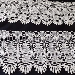 May include: A white lace trim featuring a repeating pattern of teapots and teacups arranged in vertical panels. The lace has a scalloped bottom edge and a grid-like structure, showcasing intricate thread work against a dark background.