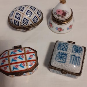 Pode incluir: Quatro pequenas caixas de porcelana para bugigangas com fechos metálicos articulados. Uma oval com grade azul e branca, uma em forma de sino com padrões florais rosa, uma retangular com grade vermelha e azul e uma quadrada com texto azul.