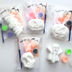 May include: Four individual clear plastic kits containing white ceramic Halloween figures including a bat, two pumpkins, and a skull. Each kit includes a paintbrush and two small paint pots. The backing cards feature the text PAINT ME.