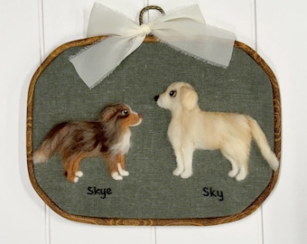 May include: An oval embroidery hoop wall hanging featuring two needle-felted dogs on olive green fabric. A brown and white dog is labeled Skye, and a cream-colored dog is labeled Sky. A white ribbon is tied at the top of the wooden frame.