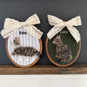 May include: Two oval-shaped cat portraits with wooden frames. One features a gray and brown cat on a blue and white striped background, labeled "Bebe." The other shows a similar cat on a green background, labeled "Cora." Both have cream-colored lace bows.
