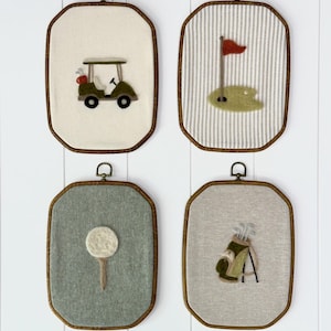 Könnte beinhalten: Vier gerahmte Wandbehänge zum Thema Golf. Jedes zeigt ein anderes gesticktes Design: einen Golfwagen, eine Fahne auf einem Grün, einen Golfball auf einem Tee und eine Golftasche mit Schlägern. Die Rahmen haben eine Holzoptik.