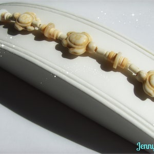 May include: A white and cream colored beaded bracelet with turtle charms. The bracelet is on a white surface.